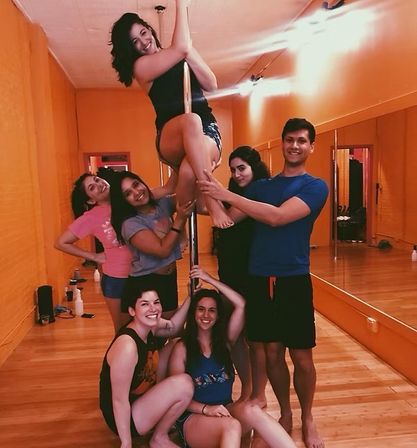 Group of friends posing around a pole in a bright orange dance studio with wood floors and mirrored wall during a pole fitness class.