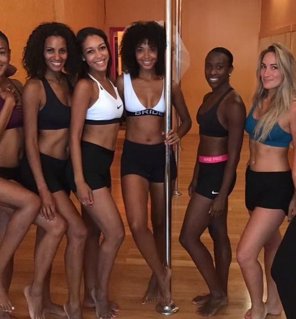 Group of women in sports bras and shorts posing barefoot around a chrome dance pole in a wood-floor pole fitness studio, smiling and ready for a workout.