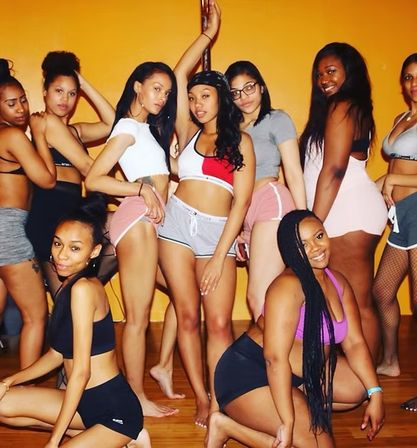 Group of young women in athletic shorts and crop tops posing around a dance pole in a bright orange fitness studio, smiling and striking confident dance poses