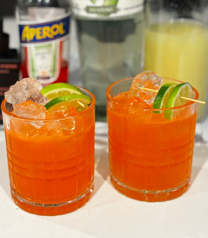 Two bright orange Aperol cocktails with ice and lime-wheel garnishes on a white countertop, blurred bottles of Aperol, tequila and juice in the background