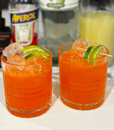 Two bright orange Aperol cocktails with ice and lime-wheel garnishes on a white countertop, blurred bottles of Aperol, tequila and juice in the background