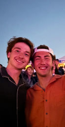 Two smiling young men posing at an outdoor evening festival or concert — one in a black shirt and jeans, the other in an orange button-up and backward white cap, with a cheering crowd and vendor tents in the background.