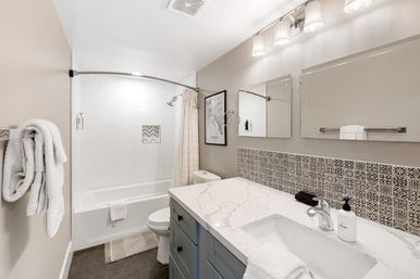 Bright modern bathroom with marble-look vanity, patterned tile backsplash, tub-shower with curved curtain rod, white towels and frameless mirrors