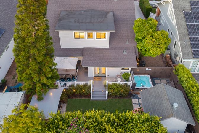 Aerial dusk view of a suburban backyard with a lit rear deck and umbrella patio, small hot tub, manicured lawn, tall evergreen tree and neighboring homes with solar panels.