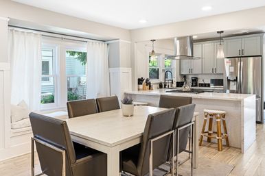 Bright open-concept dining area and modern kitchen with white island and marble countertop, stainless steel fridge and range hood, pendant lights, leather dining chairs, light wood floors and a cozy window seat