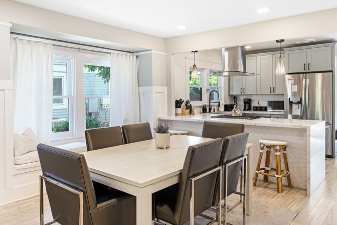 Bright open-concept dining area and modern kitchen with white island and marble countertop, stainless steel fridge and range hood, pendant lights, leather dining chairs, light wood floors and a cozy window seat