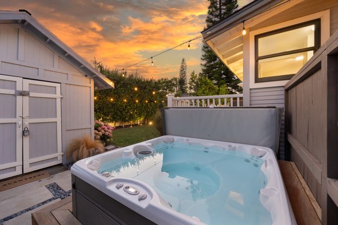 Backyard hot tub on a wooden deck at sunset with glowing string lights, manicured garden, white storage shed and privacy fence — cozy residential patio