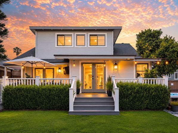 Charming modern two-story home at sunset with glowing porch lights and string bulbs, white railing, patio umbrella, French doors, and a manicured lawn with hedges.