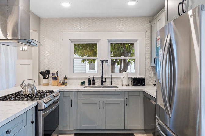 Bright modern kitchen with gray shaker cabinets, marble-look countertops, hexagon-tiled backsplash, stainless-steel refrigerator and gas range, sink beneath double window with leafy tree view