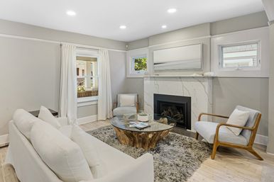 Bright modern living room interior with white sofa, two mid-century wood armchairs, marble fireplace, glass-top tree-root coffee table on a shag rug, neutral coastal-style decor and recessed lighting
