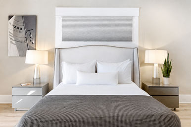 Bright modern master bedroom interior with upholstered headboard, crisp white linens, gray waffle-texture coverlet, matching bedside tables and lamps, sailboat wall art and a potted snake plant