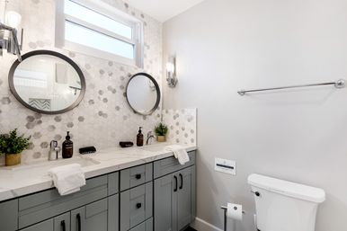 Modern spa-like bathroom with gray double-sink vanity, white marble-look countertop, hexagon mosaic tile backsplash, two round mirrors, chrome wall sconces, potted plants and toilet