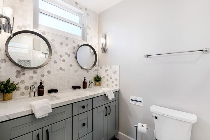 Modern spa-like bathroom with gray double-sink vanity, white marble-look countertop, hexagon mosaic tile backsplash, two round mirrors, chrome wall sconces, potted plants and toilet