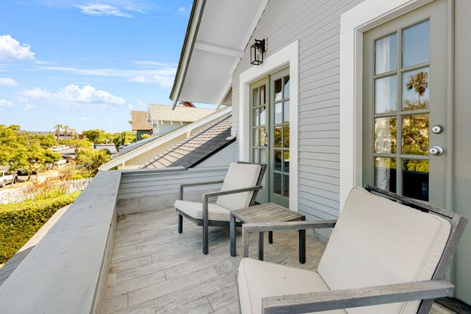 Sunny coastal balcony with two cushioned armchairs and a small wooden side table, gray siding and glass French doors overlooking a palm-lined, tree-filled neighborhood.