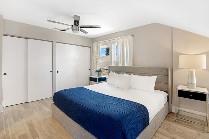 Bright modern bedroom with king bed, gray upholstered headboard, navy knit blanket, hardwood floors, ceiling fan, sliding closet doors, bedside tables and lamp