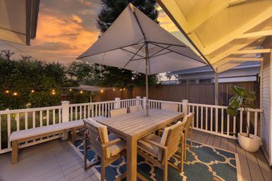 Cozy backyard deck dining with a wooden table, cushioned chairs and large umbrella, twinkling string lights and potted plant at sunset.