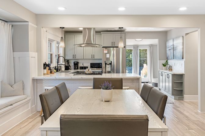 Bright open-concept modern kitchen and dining area with light wood table and leather chairs, marble-topped island with gas cooktop and stainless steel hood, pendant lights and sliding glass doors to backyard.