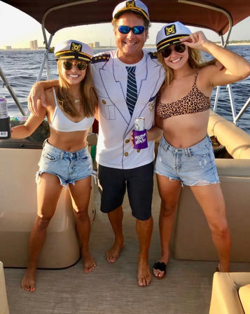 Sunlit boat party off the coast: three friends in captain hats — two women in bikinis and denim shorts and a man in a captain jacket holding canned drinks.
