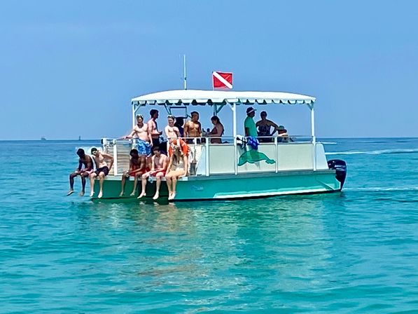 Group of people lounging on a teal pontoon boat with a diver-down flag, feet dangling over the side in clear turquoise water under a bright blue sky — sunny snorkel/boat tour vibe.