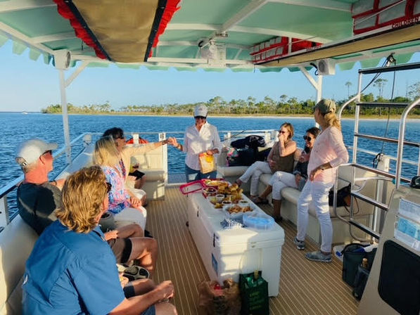 Group of adults socializing on a pontoon boat near a tree-lined shoreline, sharing snacks and drinks around a cooler-table on a sunny coastal bay — relaxed summer boat cruise.