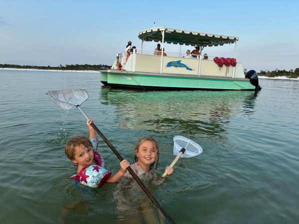 Two smiling children wading in calm shallow coastal water holding dipping nets, with a green-and-white covered pontoon tour boat and sandy shoreline in the background.