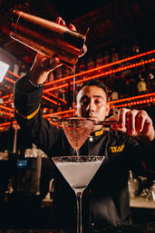 Mixologist pouring a shaken cocktail through a copper strainer into a chilled martini glass beneath red LED bar lights — stylish urban cocktail bar scene.