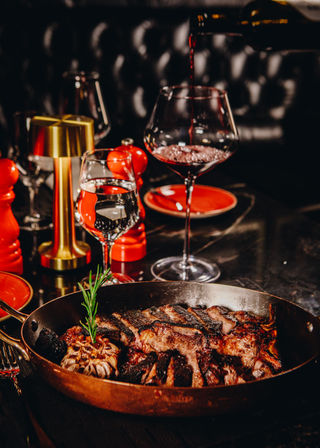 Gourmet seared ribeye steak in a copper skillet with roasted garlic and rosemary on a dim steakhouse table, red wine being poured into a glass beside water and red tableware.