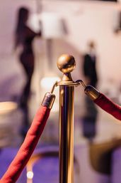 Red velvet rope clipped to a polished gold stanchion at a VIP event entrance, blurred silhouettes and moody purple lighting in the background