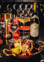 Fine-dining restaurant table with a bottle of white wine and a large wine glass, water glass, brass table lamp and red pepper mill next to a copper pan of grilled seafood with lemon wedge in a dimly lit, upscale setting.