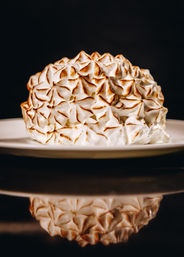Golden-tipped meringue dome (baked Alaska) on a white plate with dramatic dark background and mirrored reflection