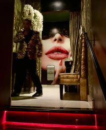 Dim, glamorous lounge interior with red LED steps, ornate gold chair and blurred person in a sequined jacket in front of a giant mural of a face with glossy red lips and a single red teardrop.