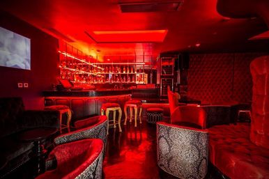 Red-lit upscale cocktail lounge interior with velvet armchairs, patterned club chairs, tufted banquettes and an illuminated back bar, moody nightlife vibe