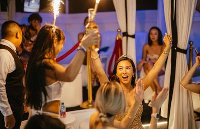 Woman wearing a tiara cheering at a VIP nightclub table as friends present bottle service with sparklers during a celebratory night out