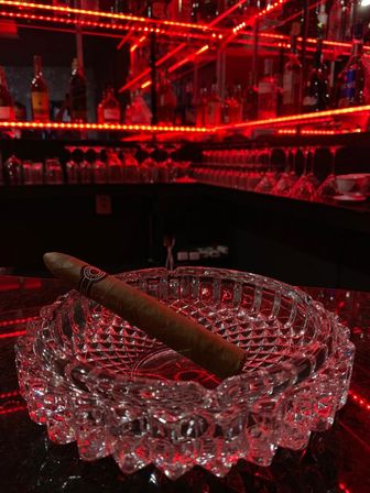 Cigar resting in a crystal ashtray on a glossy bar counter under red LED lighting, with backlit liquor bottles and hanging glassware in the background.