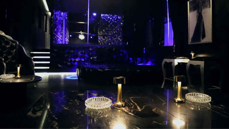 Luxurious nightclub lounge interior with purple ambient lighting, marble table in foreground featuring crystal ashtrays and brass mini lamps, tufted leather seating, raised stage and illuminated steps in the background.