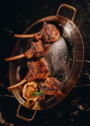 Top-down close-up of four juicy grilled lamb chops on a copper skillet with roasted garlic and a rosemary sprig, sprinkled with flaky sea salt on a dark marble surface — gourmet restaurant-style.
