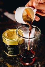 Close-up of dark espresso pouring from a white demitasse into a tall glass beside a gold-lidded jar on a dark countertop.