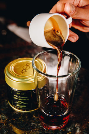Close-up of dark espresso pouring from a white demitasse into a tall glass beside a gold-lidded jar on a dark countertop.