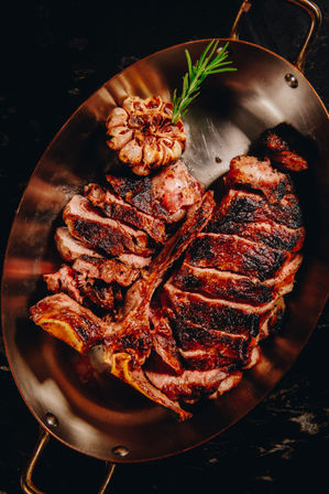 Sliced grilled tomahawk steak with charred crust and pink, juicy interior on a copper pan, garnished with roasted garlic head and a rosemary sprig — steakhouse-style presentation.