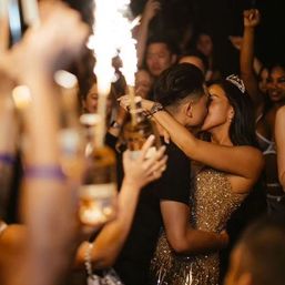 Couple kissing at a lively nightclub VIP celebration, woman in gold sequin dress and tiara embracing partner as friends cheer and raise sparklers and bottle service