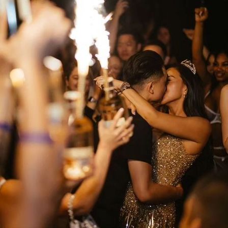 Couple kissing at a lively nightclub VIP celebration, woman in gold sequin dress and tiara embracing partner as friends cheer and raise sparklers and bottle service