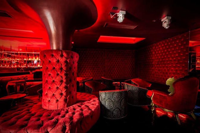 Moody red velvet lounge interior with a tufted circular banquette around a central column, ornate armchairs, patterned club chairs, low tables and a backlit bar — luxury nightclub or cocktail lounge vibe.