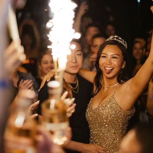 Smiling woman in a sparkly gold dress and tiara raises her arms at a lively nightclub celebration with friends, sparklers and champagne bottles.