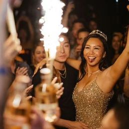 Smiling woman in a sparkly gold dress and tiara raises her arms at a lively nightclub celebration with friends, sparklers and champagne bottles.