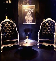 Moody lounge scene with two ornate tufted black leather throne chairs, round marble coffee table with a candle, and a framed black-and-white vintage Hollywood pinup poster on a dark wall.