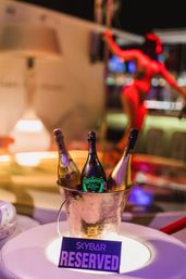 Champagne bucket with three bottles and a "Reserved" sign on a lit VIP table in a colorful nightclub lounge, blurred dancer silhouette in the background.