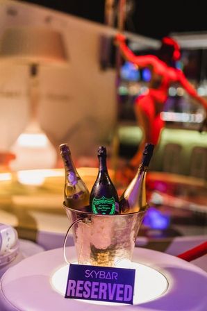 Champagne bucket with three bottles and a "Reserved" sign on a lit VIP table in a colorful nightclub lounge, blurred dancer silhouette in the background.