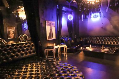 VIP nightclub lounge interior with tufted black leather sofas, low cocktail tables, purple mood lighting, chandeliers, and vintage framed posters