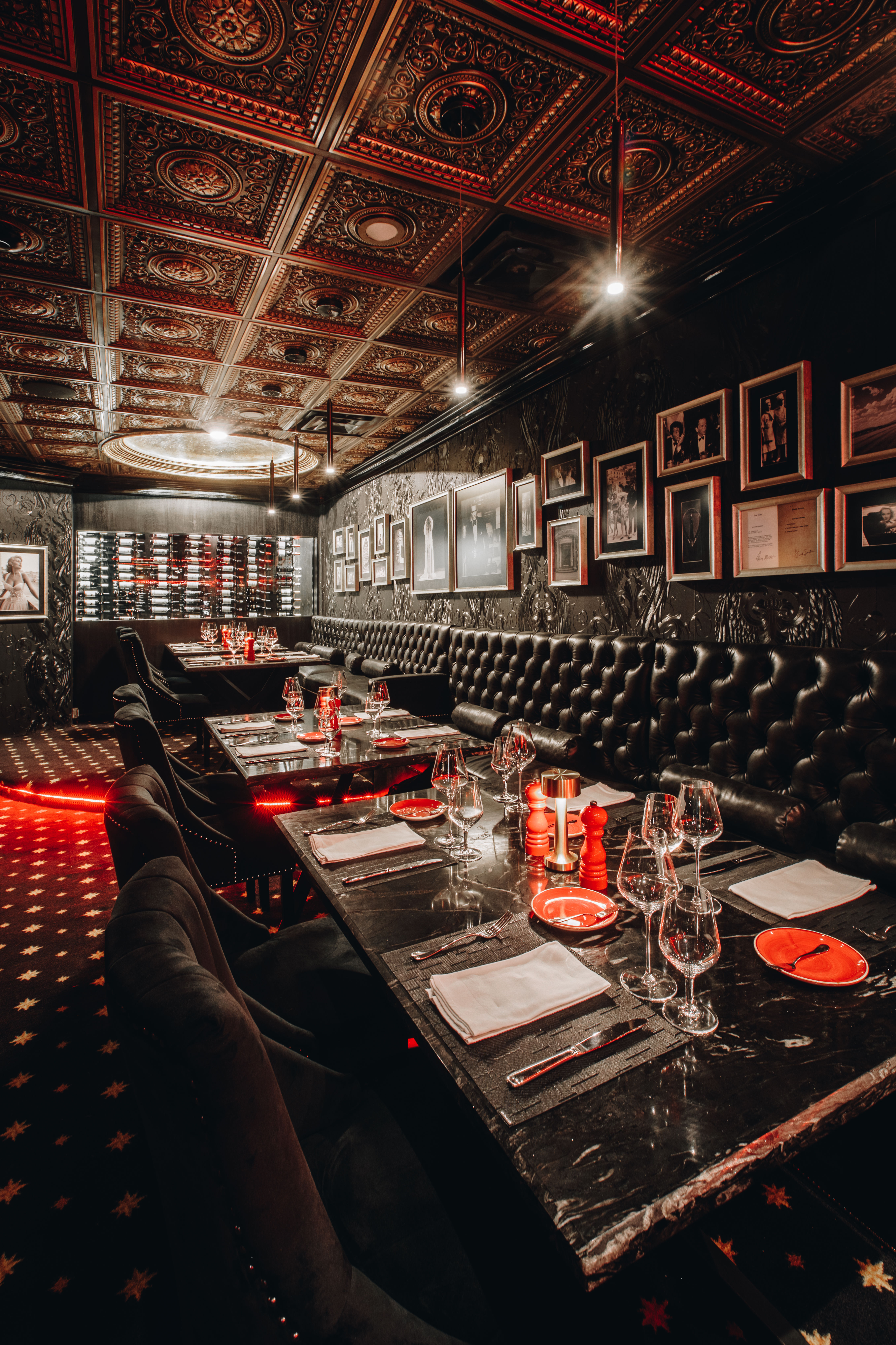 Moody, glamorous dining room in an upscale restaurant with tufted black leather banquettes, marble tables set with glassware and red accents, ornate embossed coffered ceiling, framed photos on dark walls and a backlit wine wall.