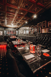 Moody, glamorous dining room in an upscale restaurant with tufted black leather banquettes, marble tables set with glassware and red accents, ornate embossed coffered ceiling, framed photos on dark walls and a backlit wine wall.
